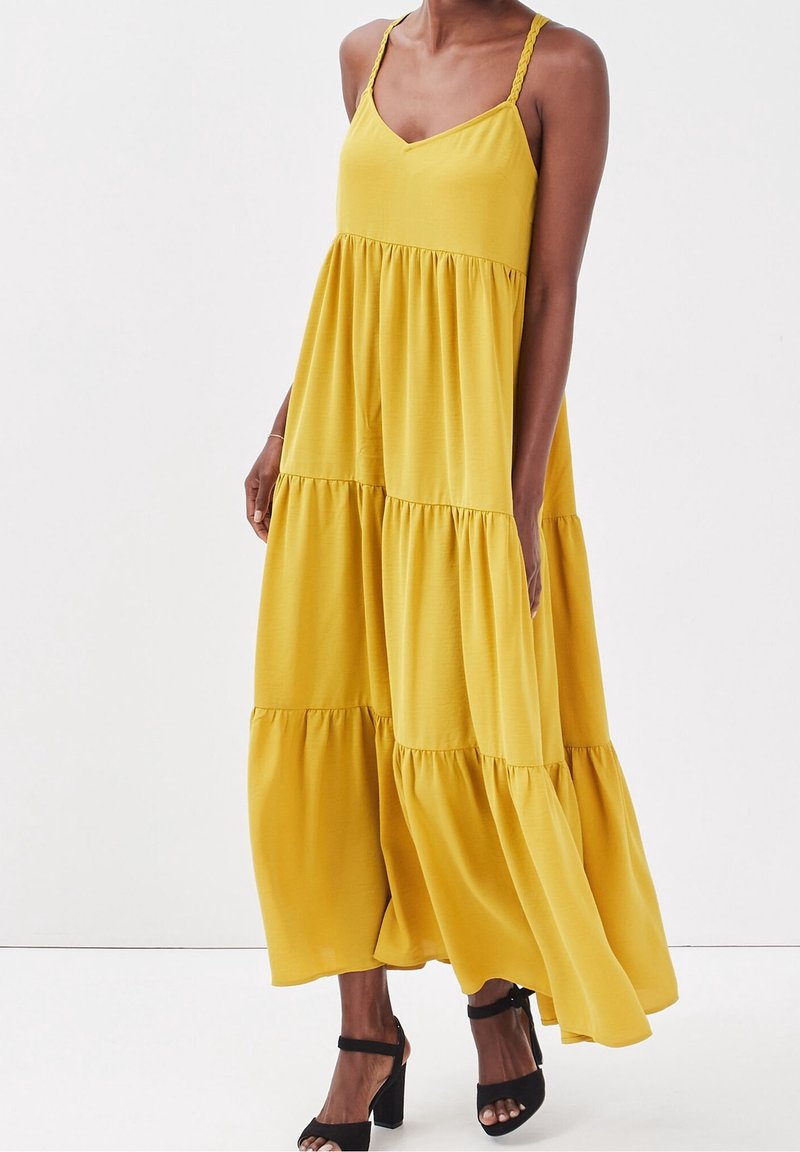 Femme portant une robe maxi jaune sans manches à volants avec des bretelles tressées et des sandales à talons hauts noires, debout devant un fond uni.