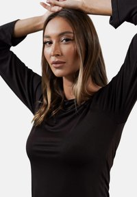 Czarna bluzka z długim rękawem i opadającym dekoltem. Gładki, elastyczny materiał, lekko dopasowany krój. Modelka pozuje z uniesionymi rękami na białym tle.