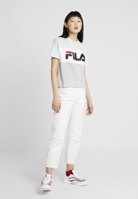 T-shirt Fila en bleu clair et gris avec logo, associé à un jean cropped blanc. Le modèle porte des baskets rouges et blanches.