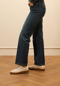 Dunkelblaue, hoch taillierte, gerade geschnittene Jeans mit einem subtilen Wasch-Effekt. Kombiniert mit weißen Sneakern mit strukturierten Akzenten und einer Gummisohle.