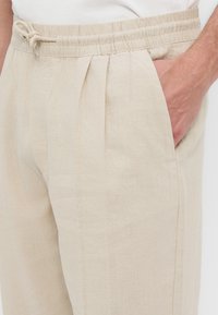Pantalon en lin beige clair avec taille élastique et cordon, poches latérales, et main placée dans une poche.