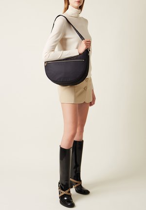 Borsa a spalla nera con texture in forma di mezzaluna, con zip dorata e una lunga tracolla regolabile. Abbinata a una maglietta crema e pantaloni corti beige.