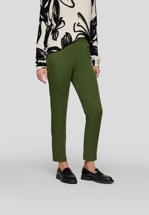 Grüne Hose im Slim-Fit, aus Baumwolle, mit Fronttaschen und glatter Textur. Kombiniert mit schwarzen Slippern und einem gemusterten Pullover.