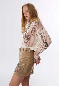 Strickpullover mit lockerer Passform, ausgestattet mit einem abstrakten Muster in Beige und Braun. Wildleder-Minirock in hellbeige mit Metallnieten und einem Gürtel.