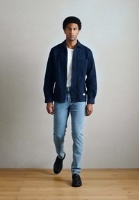 Model má na sebe tmavomodrú košeľu na gombíky s vreckom, biele tričko, svetlomodré džínsy a čierne topánky, stojí na drevenej podlahe.