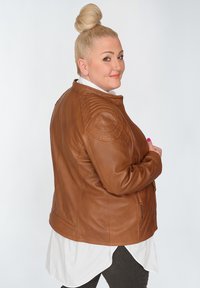 Braune Leder-Motorradjacke mit gesteppten Schulterakzenten, Reißverschlusstaschen und taillierter Silhouette, getragen über einem weißen Hemd.