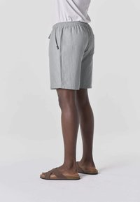 Persona vistiendo pantalones cortos casuales de color gris claro y sandalias marrones sin talón, de pie sobre un fondo blanco liso, de perfil.