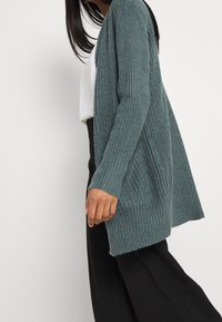 Tyrkysový žebrovaný kardigan s volným střihem, dlouhými rukávy a otevřeným předním designem. Vyrobený z měkkého pleteného materiálu s texturovaným povrchem.