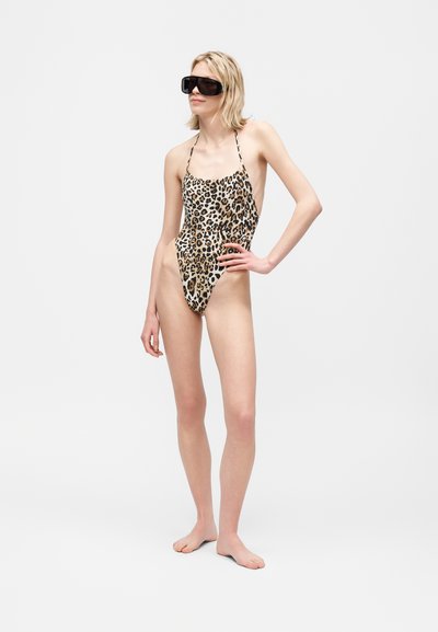 Γυναίκα που φοράει ολόσωμο μαγιό με σχέδιο λεοπάρ και μεγάλα μαύρα γυαλιά ηλίου, όρθια ξυπόλυτη με το ένα χέρι στη μέση της μπροστά σε απλό φόντο.