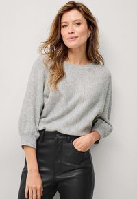 Pull en tricot gris clair avec un col rond et une silhouette décontractée, associé à un pantalon noir ajusté. Tissu texturé visible.