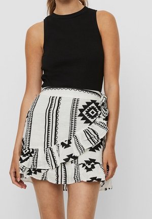 Top sans manches noir à côtes associé à une jupe blanche ornée d'un motif géométrique et tribal noir, avec un ourlet volanté et un détail de lien sur le côté.