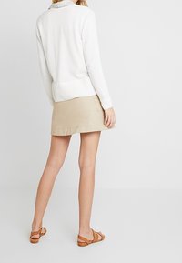 Maglione bianco con orlo a coste, abbinato a una gonna beige. Sandali marroni completano l'outfit. Vista da dietro, che mette in mostra il design a strati.