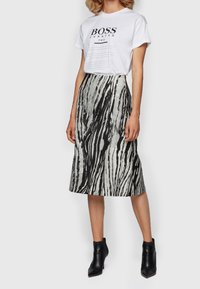 Femme portant un t-shirt graphique blanc, une jupe midi à motifs noirs et blancs, et des bottes noires à la cheville, se tenant devant un fond uni.