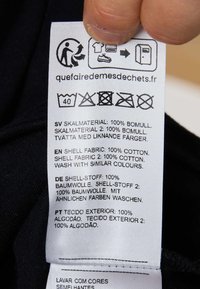 White fabric care label attached to a black garment, featuring multilingual washing instructions, care symbols, and recycling icons.