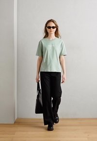 Svetlozelené tričko s krátkymi rukávmi a čierne široké nohavice. Čierne slnečné okuliare a čierna taška cez rameno dopĺňajú outfit. Pozadie z drevenej podlahy.