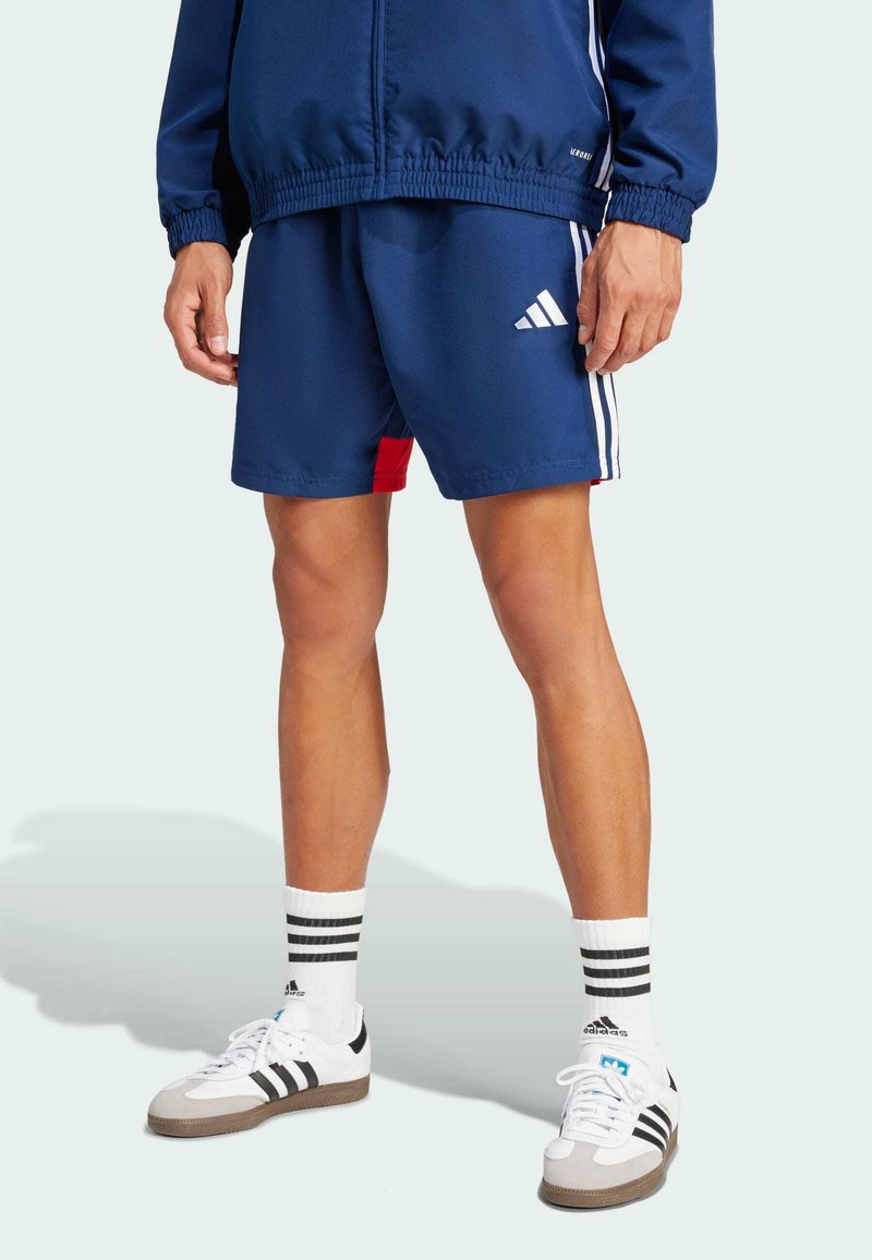 Blue athletic shorts with red accent, white three-stripe detail, paired with white shoes featuring black stripes and textured rubber soles.