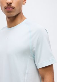 Κοντινό πλάνο ενός ατόμου που φοράει ανοιχτό μπλε αθλητικό μπλουζάκι με κοντά μανίκια και ένα μικρό λευκό λογότυπο στο στήθος, με απλό φόντο.