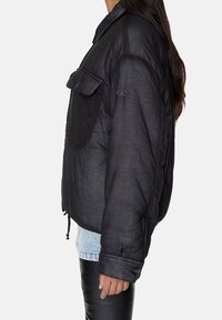 Czarna pikowana kurtka o nowoczesnym kroju, z przednią kieszenią, kontrastującymi przeszyciami oraz elastycznymi mankietami. Tkanina wydaje się gładka i teksturowana.