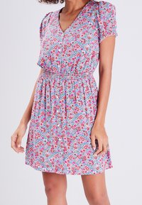 Robe à imprimé floral avec un décolleté en V profond, des manches bouffantes courtes et une taille élastique. Les couleurs incluent le rose, le rouge et le bleu sur un fond clair.