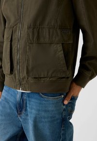 Bomberka w kolorze oliwkowej zieleni wykonana z lekkiego materiału, z dwoma przednimi kieszeniami i ściągaczami na mankietach, zestawiona z niebieskimi dżinsami.