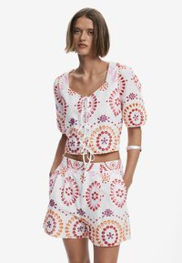 Vit kort topp med lila, orange och röda blommönster, med V-ringning och dragsko. Matchas med kortbyxor och sidofickor.