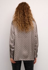 Camisa de manga larga en satén gris con puntos negros. Presenta un corte recto, puños abotonados y posibles aberturas laterales en el dobladillo.