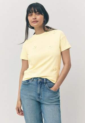 T-shirt in cotone giallo con maniche corte, caratterizzata da fiori applicati sul davanti, abbinata a jeans in denim blu chiaro. Design semplice.