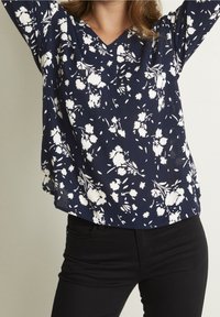 Femme portant un chemisier bleu marine avec des motifs floraux blancs et un pantalon noir, levant les bras au-dessus de sa tête sur un fond uni.