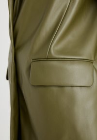 Olijfgroen lederjack met een gladde textuur, voorzien van een rechte zak en hoekige naaddetails voor een getailleerde uitstraling.