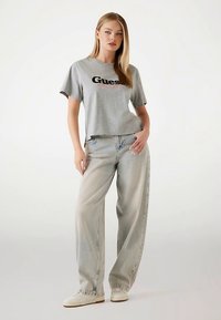 T-shirt gris court avec logo "Guess" noir. Associé à un jean ample en denim clair. Le modèle porte des baskets blanches. Fond uni.