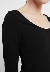 Kvinna som bär en åtsittande svart långärmad tröja med en bred rundad halsringning, visad från axel till mitten av överkroppen mot en enkel bakgrund.