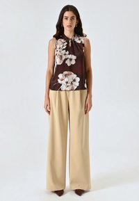 Donna che indossa un top floreale senza maniche di colore marrone scuro, pantaloni beige a gamba larga e scarpe a punta marroni, in piedi contro uno sfondo chiaro e uniforme.
