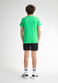Groene katoenen t-shirt, korte mouwen, klassieke ronde hals, gecombineerd met zwarte shorts. Model draagt witte en zwarte sneakers met gestreepte sokken.