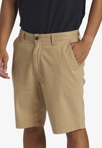 Pantalones cortos de algodón beige con un corte relajado, bolsillos estándar y cierre de botón. La tela tiene una textura suave y un detalle de costura mínimo.
