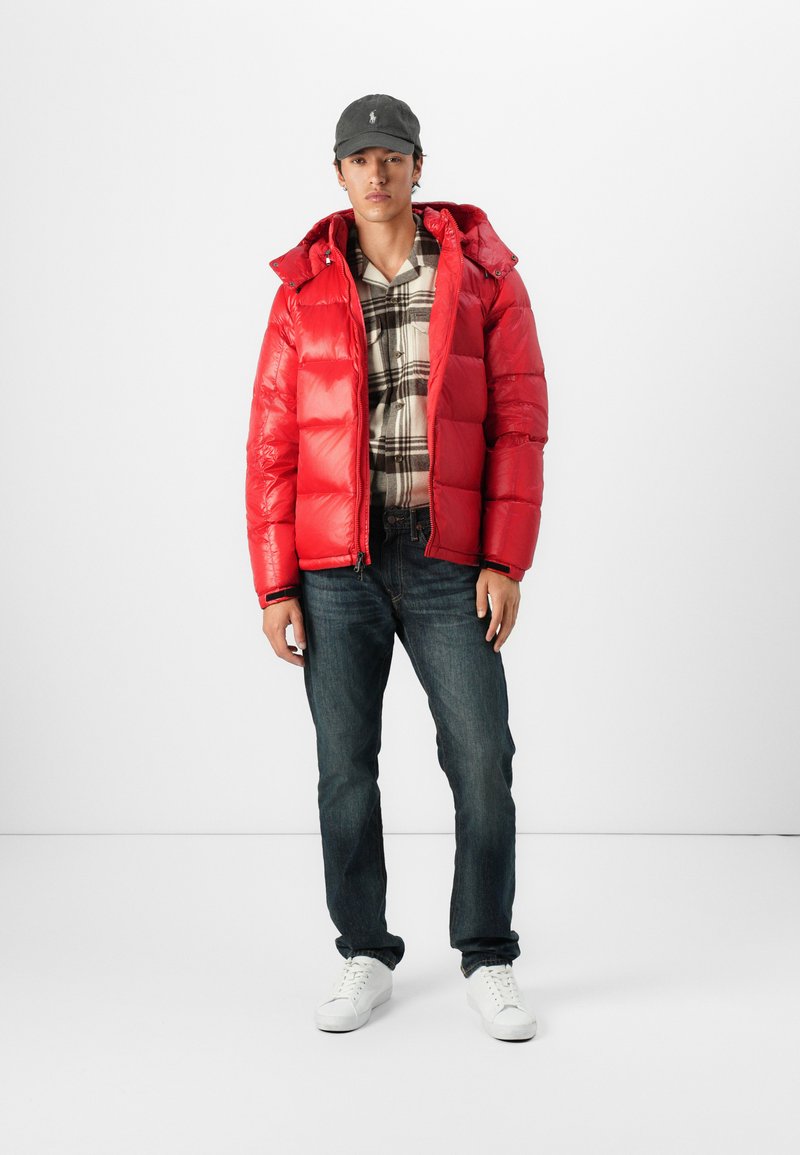Rote Pufferjacke mit glänzender Oberfläche, Kapuze und elastischen Bündchen, kombiniert mit einem karierten Hemd, dunklen Jeans und weißen Sneakers.