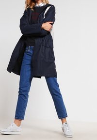 Navy waterproof parka features a hood, front pockets, and a contrasting light gray panel along the shoulders; paired with blue jeans and white sneakers.