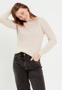 Pull beige tricoté avec texture côtelée, décolleté rond et trois boutons décoratifs sur l'épaule, accompagné de jeans noirs taille haute.