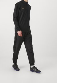Schwarzes Langarmpolo mit halbem Reißverschluss, kombiniert mit schwarzen Jogginghosen und schwarzen Fußballschuhen mit blauen Akzenten; Nike-Logo auf der Brust und den Schuhen.