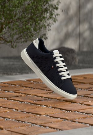 Μαύρο πλεκτό αθλητικό παπούτσι με λευκή σόλα και κορδόνια που αιωρείται πάνω από ξύλινο και πετρόστρωτο έδαφος σε εξωτερικό χώρο.