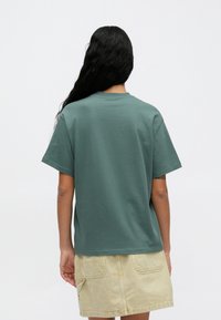 Πράσινο βαμβακερό μπλουζάκι με χαλαρή εφαρμογή, στρογγυλή λαιμόκοψη και κοντά μανίκια, συνδυασμένο με μια μπεζ φούστα που διαθέτει δύο πίσω τσέπες.