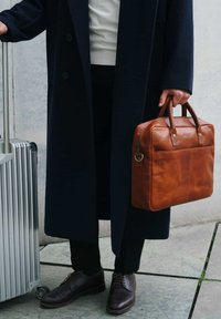 Brown leather briefcase with two handles, featuring a front pocket and zipper. Accompanied by a silver rolling suitcase on a stone surface.