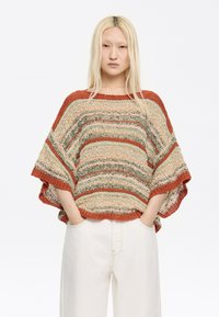 Strickponcho in erdigen Tönen von Orange, Grün und Creme mit weitem, lockeren Schnitt. Besitzt offene Seiten und gerippte Kanten. Kombiniert mit weißen Hosen.