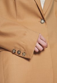 Hand partially inside pocket of tan blazer with four buttons on sleeve and one button fastened on front.