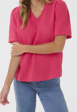 Kobieta w różowej, teksturowanej bluzce z krótkim rękawem i koronkowymi wykończeniami oraz w jasnoniebieskich dżinsach z wysokim stanem, z założonymi ramionami na torsie.