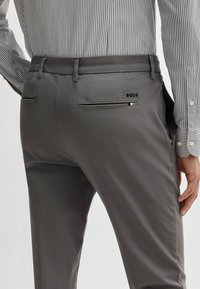 Pantalons gris ajustés en tissu lisse, dotés de deux poches arrière et d'un petit logo noir sur la poche droite. Chemise rayée visible au-dessus.