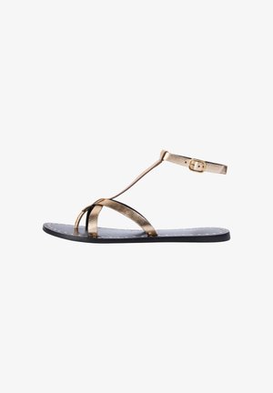 Kulta metallinen sandaali ohuilla remmeillä, varvaslenkillä ja nilkkahihnalla. Mustassa pohjassa on pieniä kultaisia niittejä tekstuurin ja yksityiskohtien tuomiseksi.