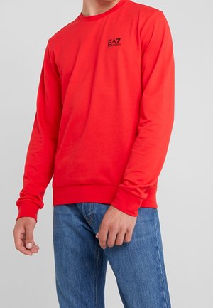 Homme portant un sweat-shirt rouge vif à manches longues avec le logo EA7 Emporio Armani sur la poitrine et un jean bleu sur un fond uni.