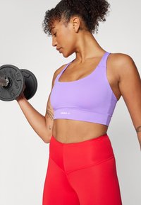 Sostén deportivo morado con un diseño de tirante ancho, combinado con leggins rojos de talle alto. Incluye el logotipo de la marca en la parte frontal. Textura de tela plana.
