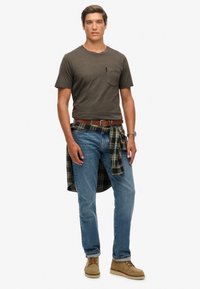 Szara koszulka z krótkim rękawem z kieszonką, niebieskie dżinsy, koszula w kratę związana w talii, brązowy pasek, beżowe buty o teksturowanej powierzchni.