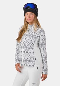 Polarowa kurtka o białej bazie z szarymi wzorami geometrycznymi i płatkami śniegu, z zamkiem z przodu, wysokim kołnierzem, w połączeniu z białymi spodniami i goglami narciarskimi.